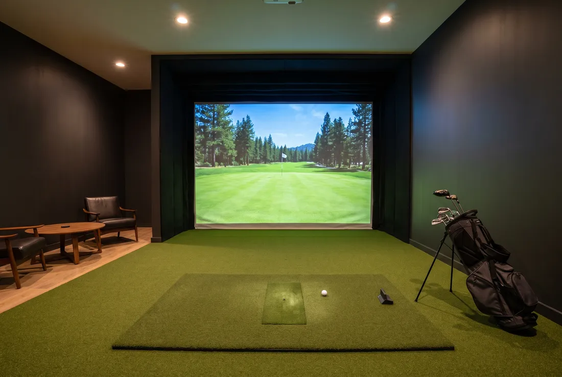 An empty indoor golf simulator bay: a large screen showing a green fairway between pines, a turf hitting mat with a ball on a tee, a golf bag and two lounge chairs.