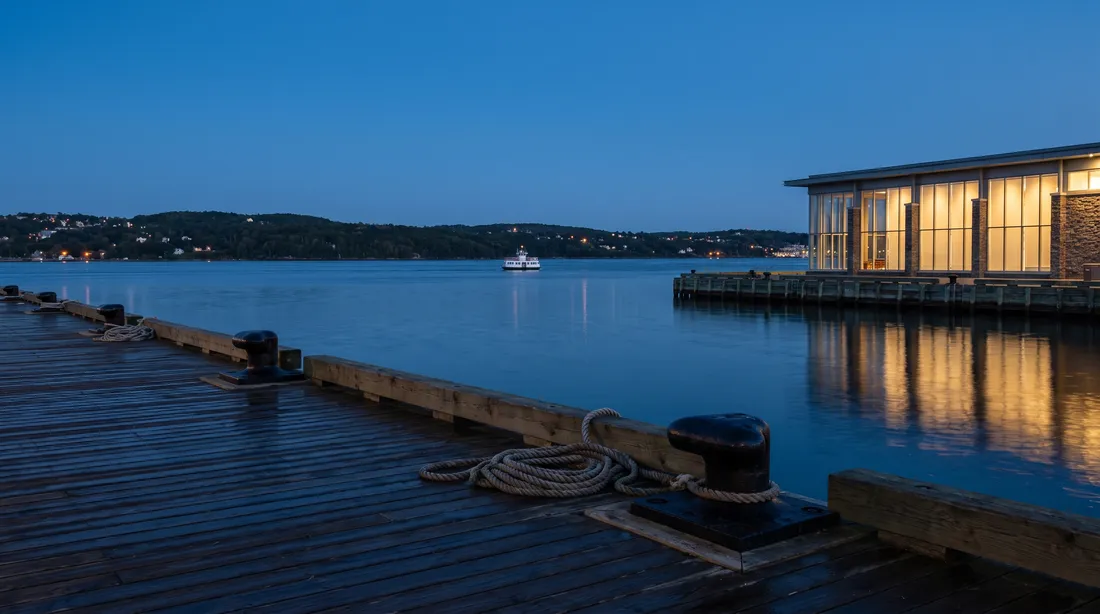 A wooden harbour boardwalk at blue hour with iron bollards and coiled rope, calm water and a lit glass building on the right.