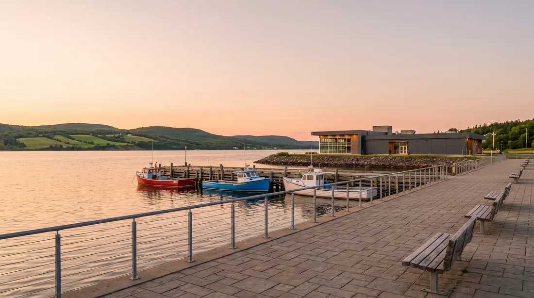 A paved harbourfront walkway with a steel railing and benches at sunset, small fishing boats at a wooden wharf and green hills across the water.